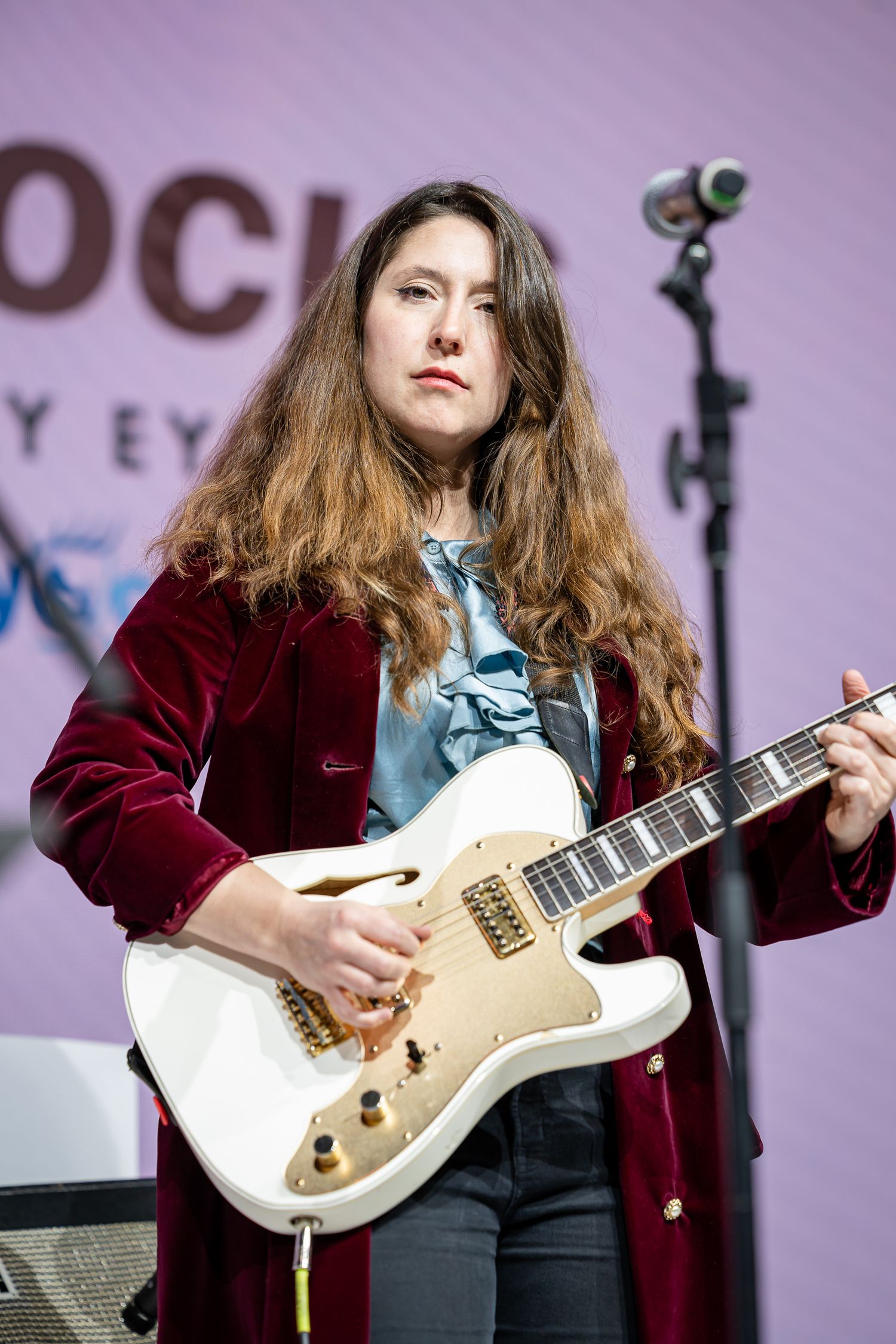 Musician performs on white electric guitar wearing burgundy velvet jacket on stage.