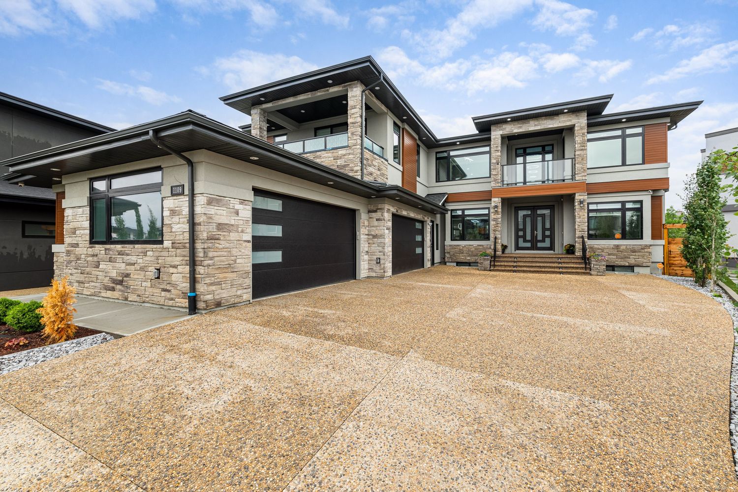 Modern two-story home with stone facade, triple garage, and exposed aggregate driveway under a cloudy sky.