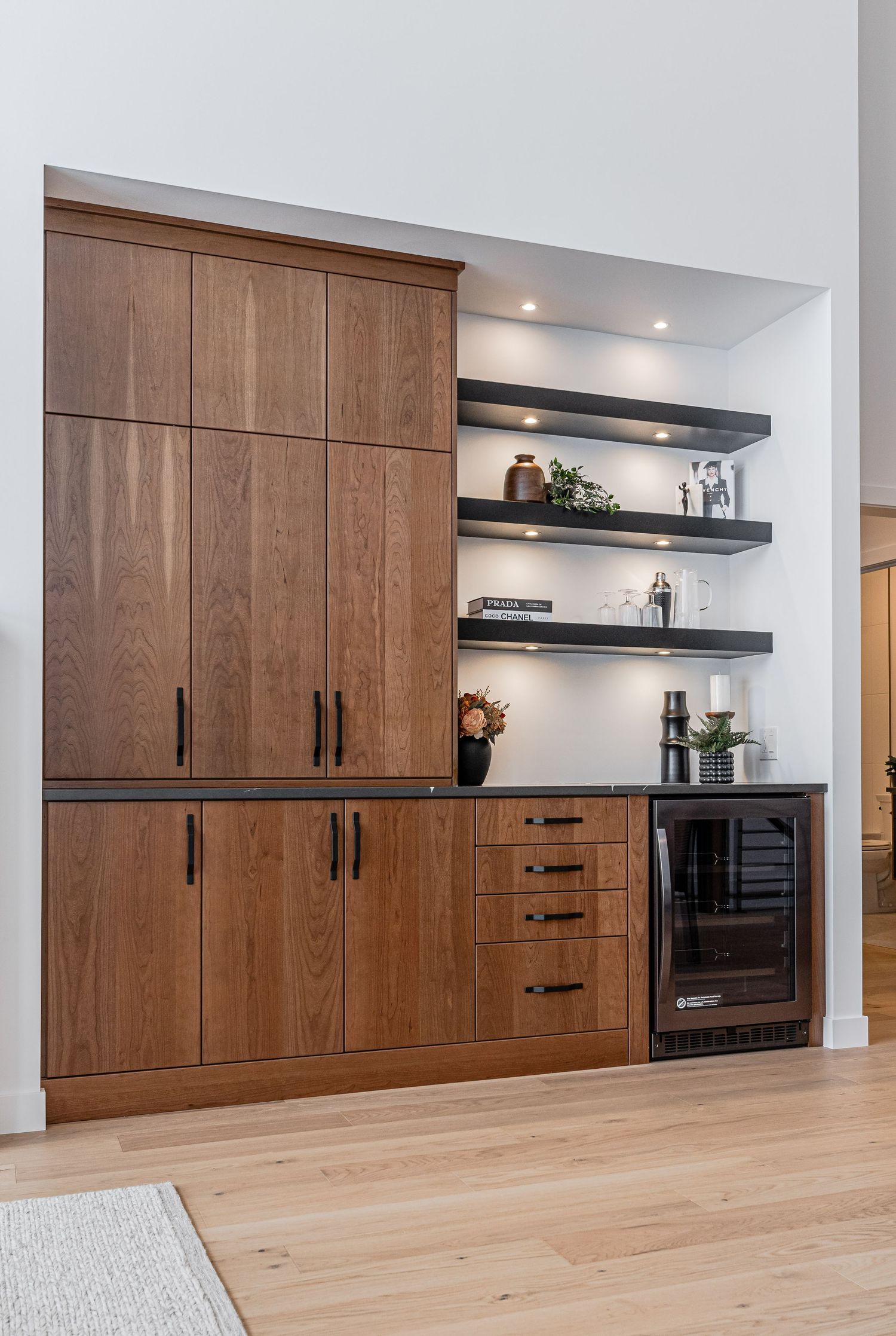 Modern built-in bar cabinet with warm wood finish, black hardware, open shelving, and wine fridge in elegant home interior.