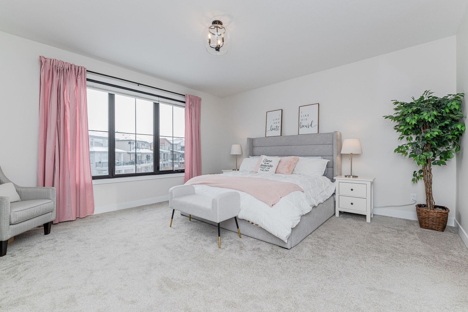 Bright bedroom with gray upholstered bed, pink curtains, carpet flooring, white nightstands, and a decorative potted tree.