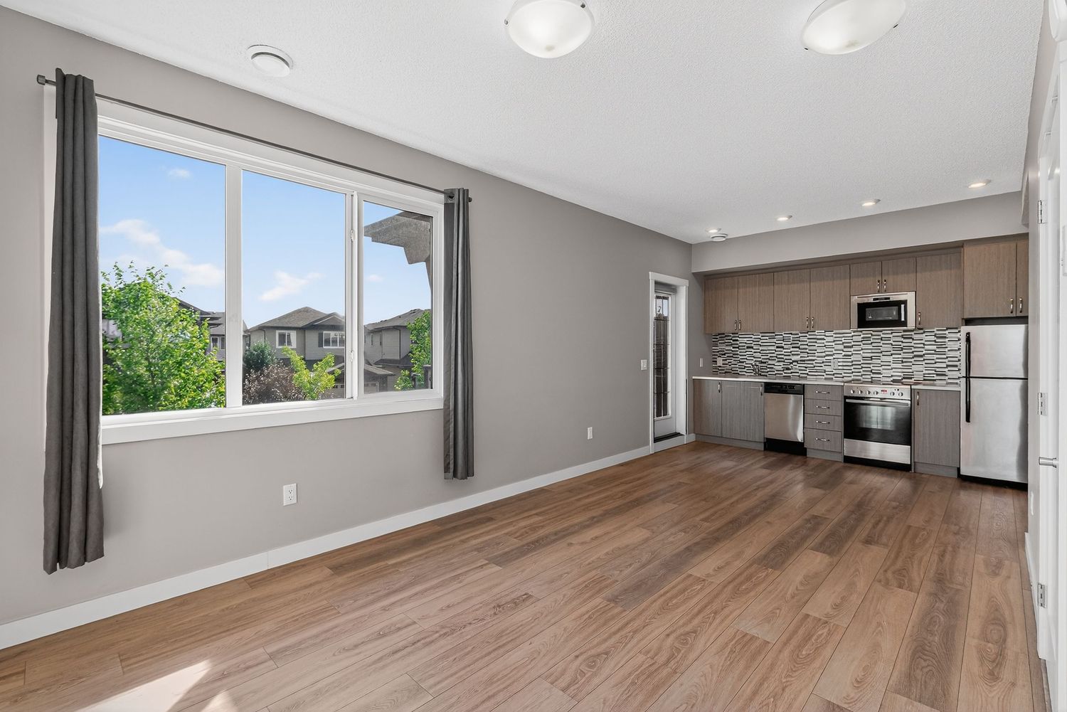 Modern open-concept kitchen and living area with hardwood floors, gray cabinets, stainless steel appliances, and large window.