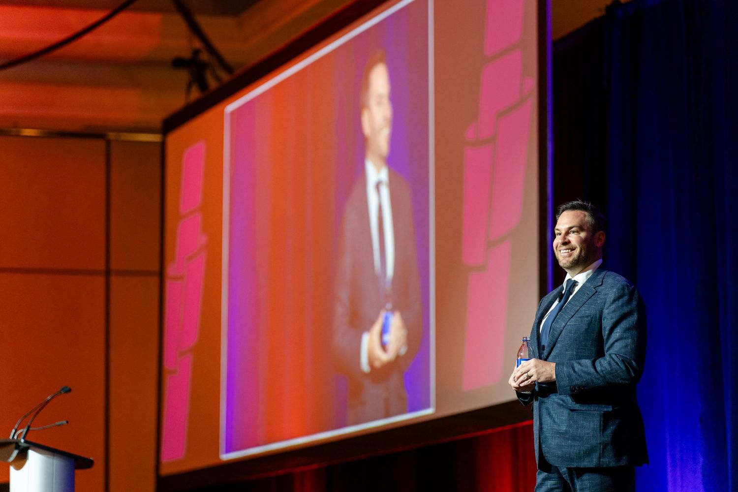Speaker presents on stage with projected presentation screen in background at professional conference.