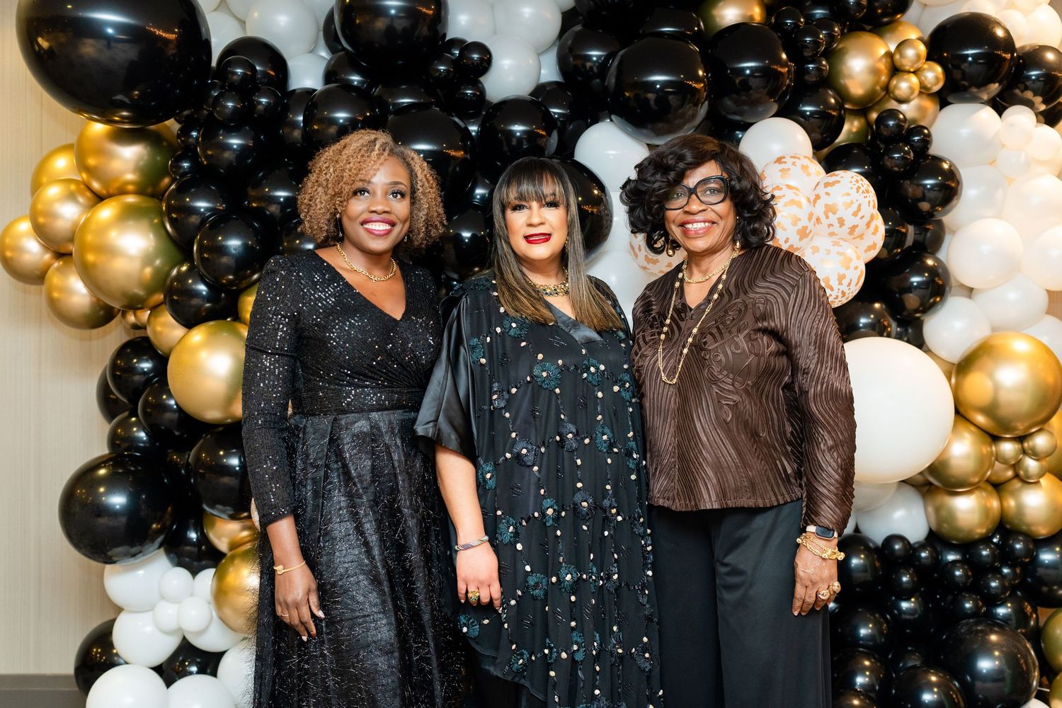 Stylish attendees pose together against a backdrop of black and gold balloons at a formal celebration.