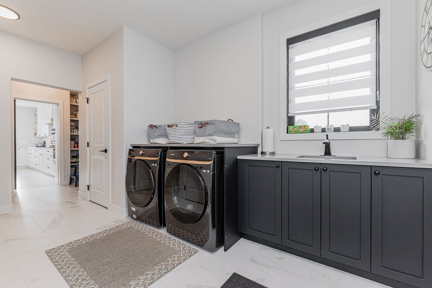 Modern laundry room with dark cabinets, front-load washer and dryer, sink, and bright natural light from a window.