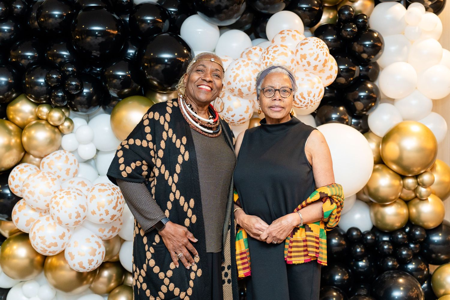 Two attendees pose together against elegant black and gold balloon wall at formal event.