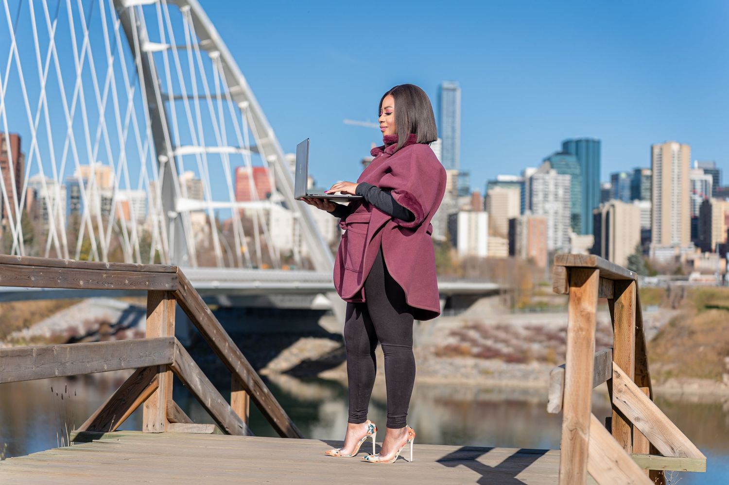 Professional woman in pink coat standing on bridge with city skyline in background.