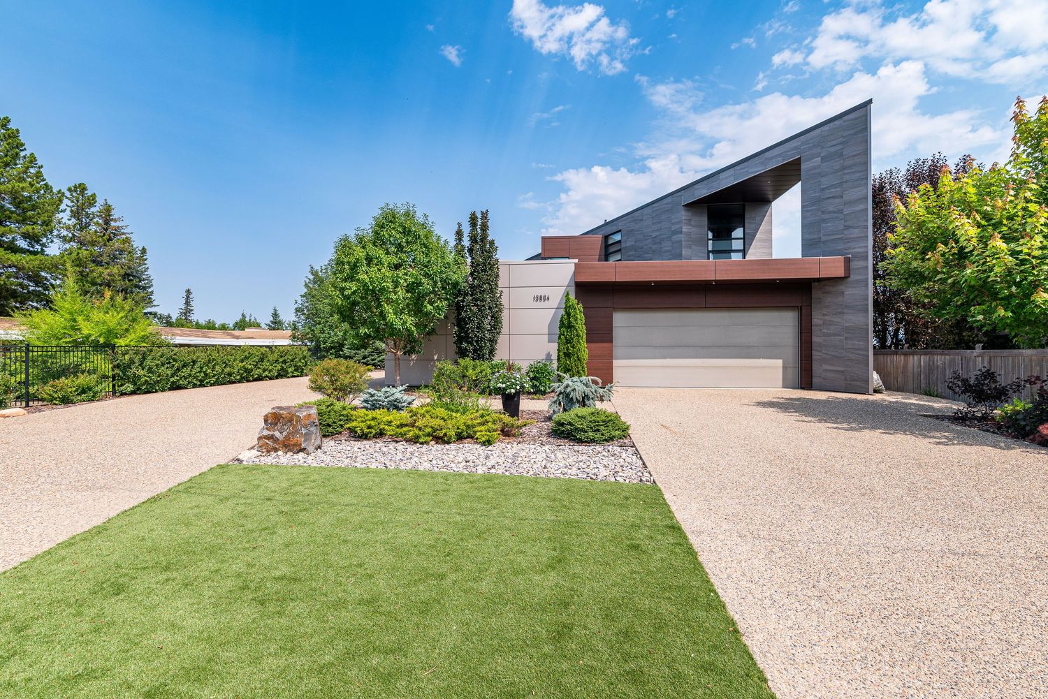 Modern angular home with gray stone facade, wooden garage, gravel driveway, and manicured green lawn surrounded by lush trees.