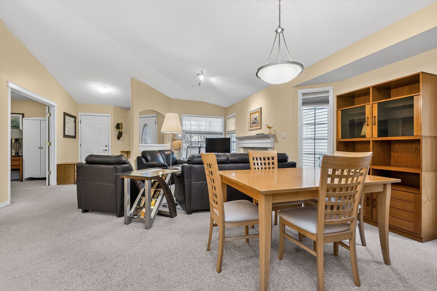 Cozy dining area with wooden table and chairs, open to a carpeted living room with leather sofas and a hutch.