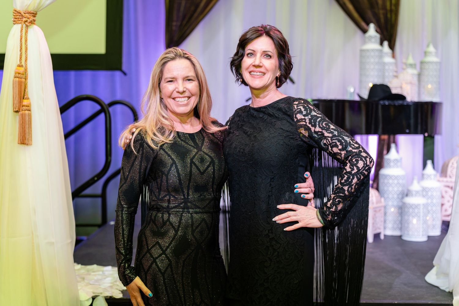 Two women in black lace dresses pose together at an elegant event venue.
