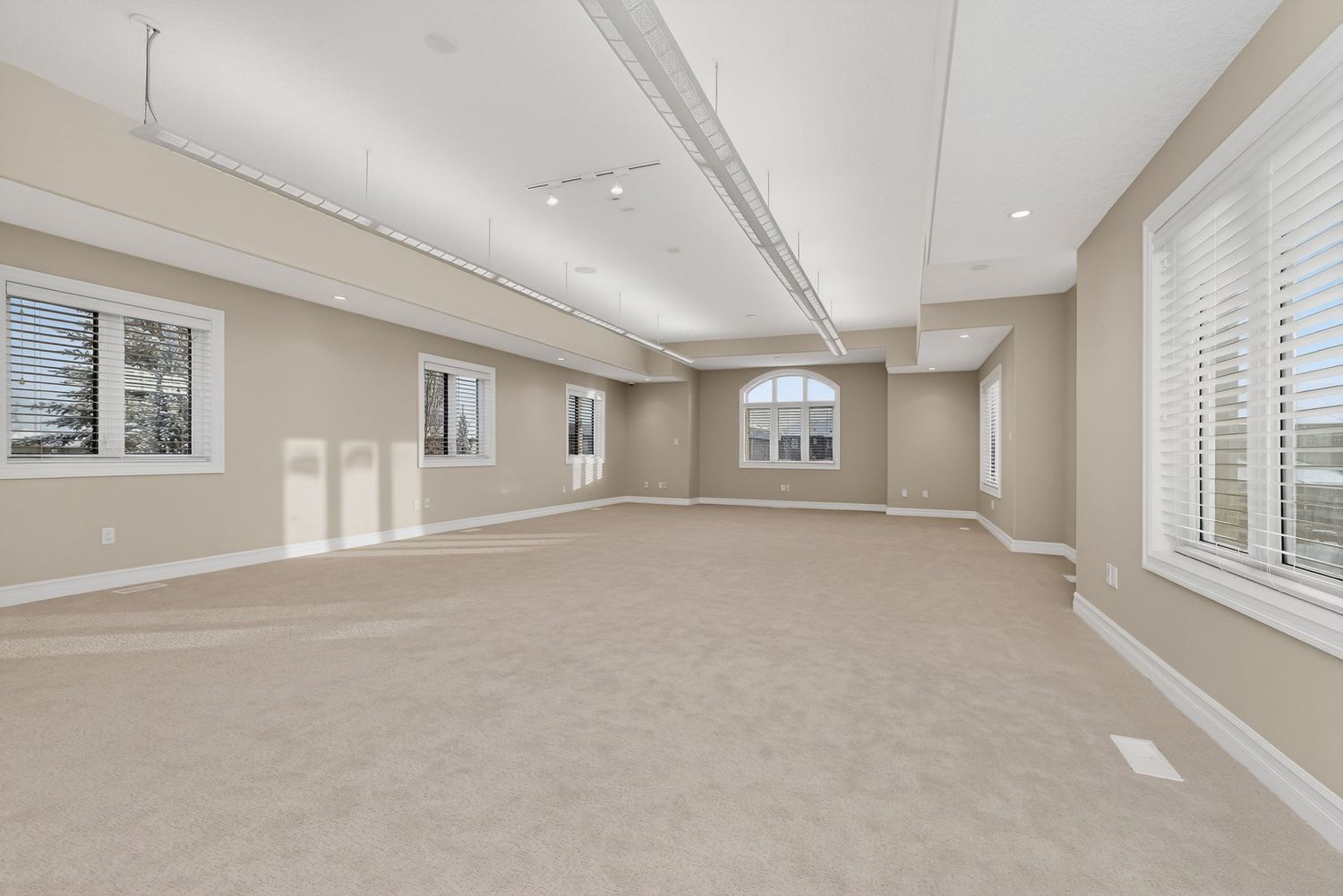 Spacious empty room with beige carpet, neutral walls, track lighting, and large windows allowing natural light.