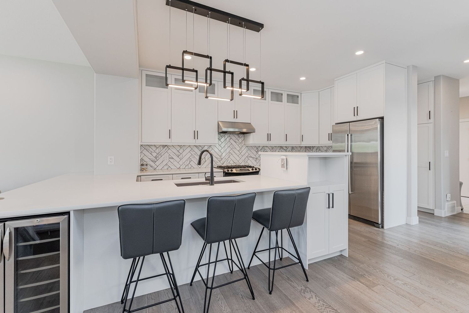 Modern white kitchen with quartz island, gray bar stools, pendant lights, herringbone backsplash, and stainless steel appliances.