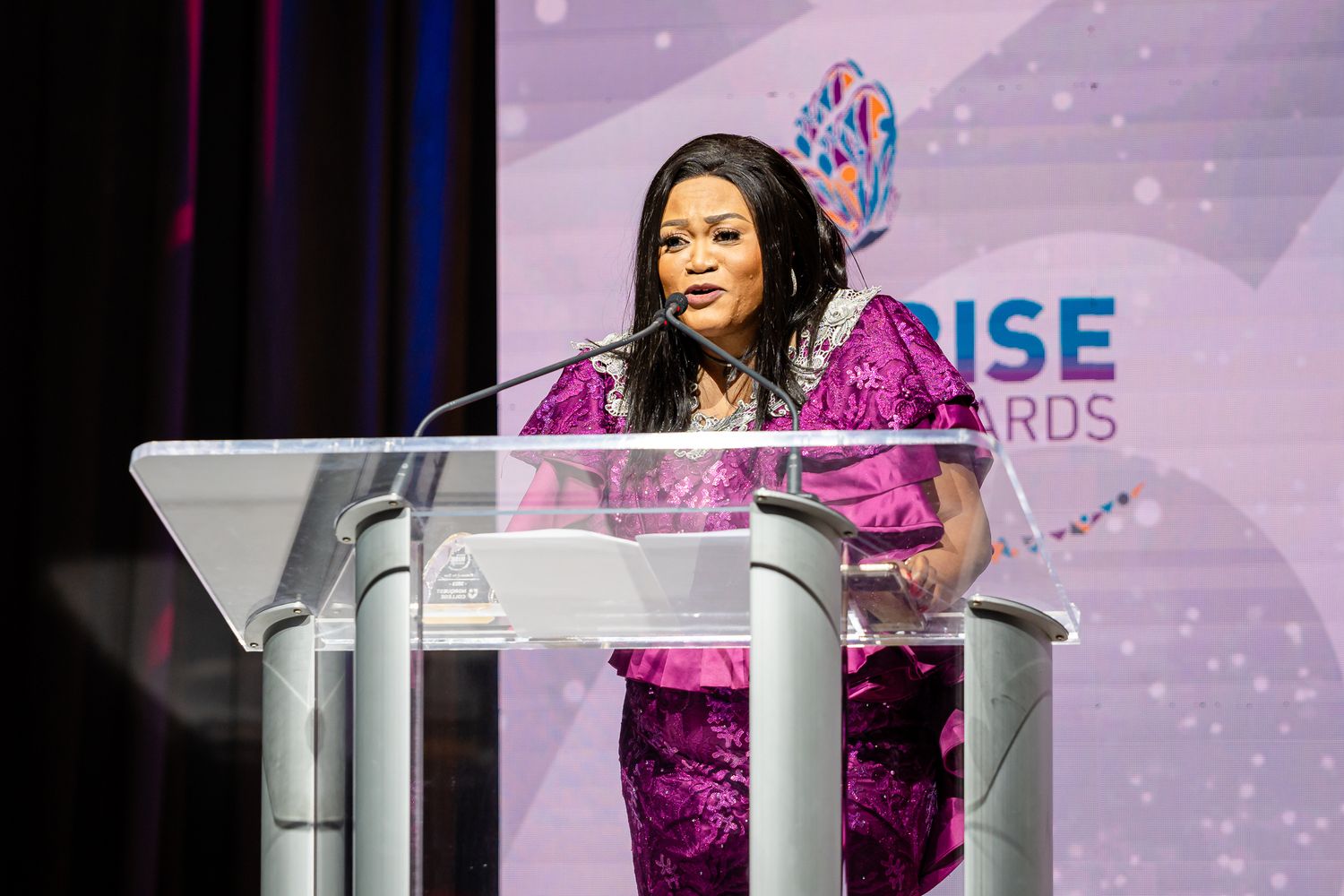 Speaker presents from white podium at RISE Awards with purple lighting and branded backdrop.