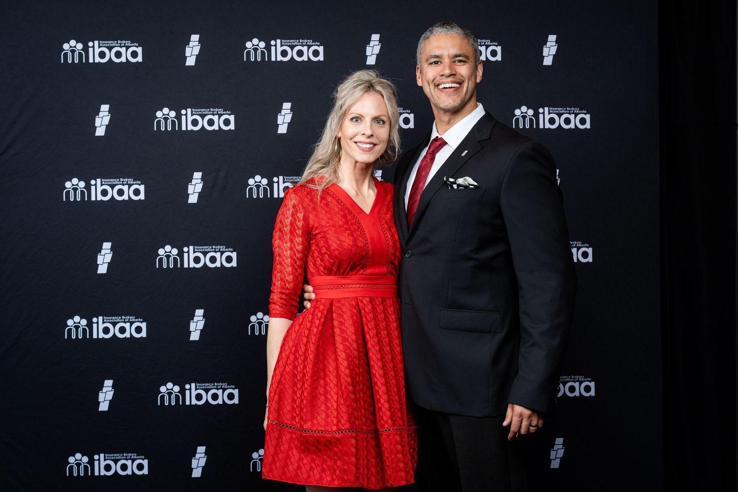 A couple poses together at an IBAA event, with the woman wearing a red dress and the man in a dark suit.