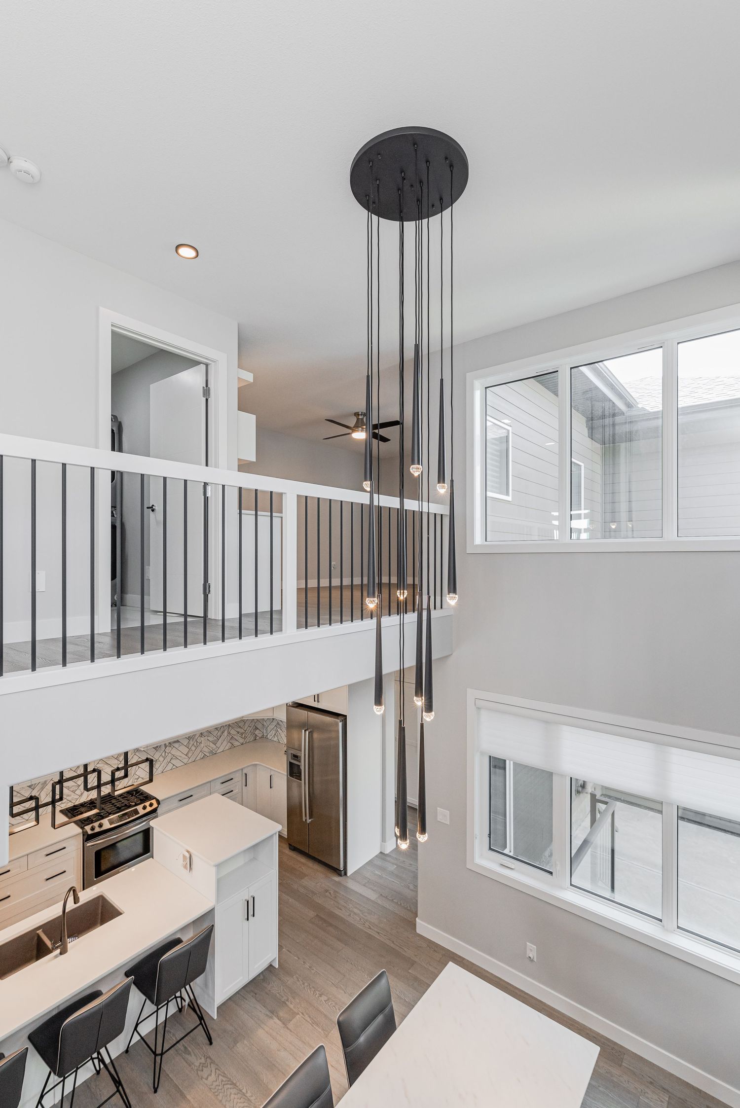 Modern open-concept home with double-height ceilings, pendant lighting, white kitchen, and upper loft railing overlooking living area.