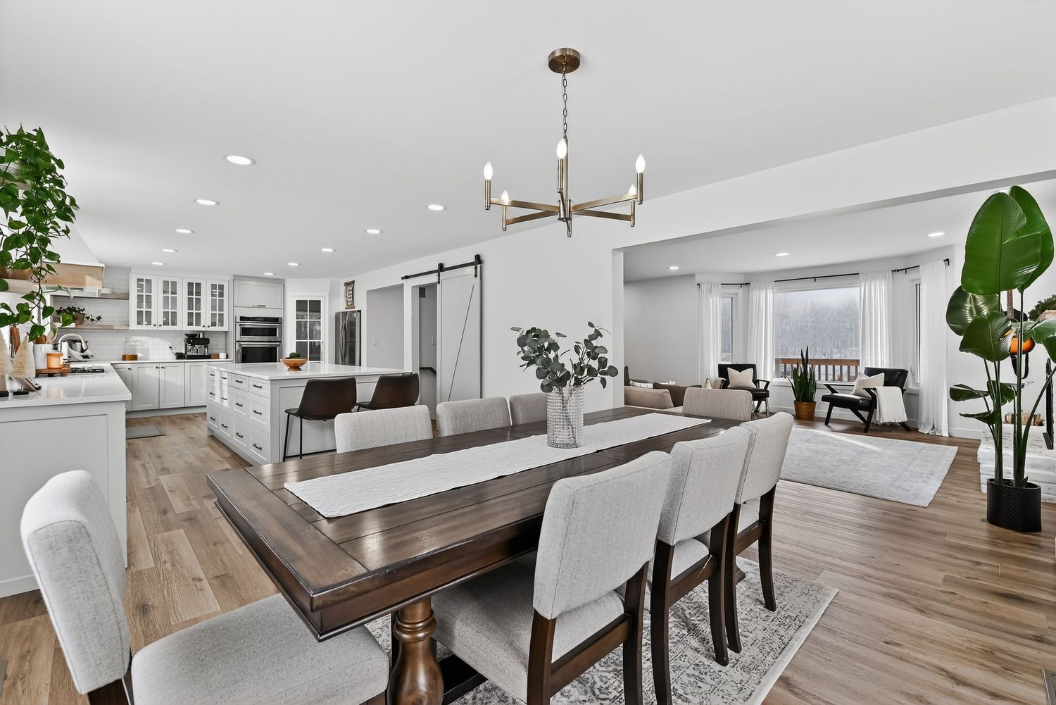 Modern open-concept dining room with wood table, white chairs, hardwood floors, chandelier, and barn door leading to kitchen.