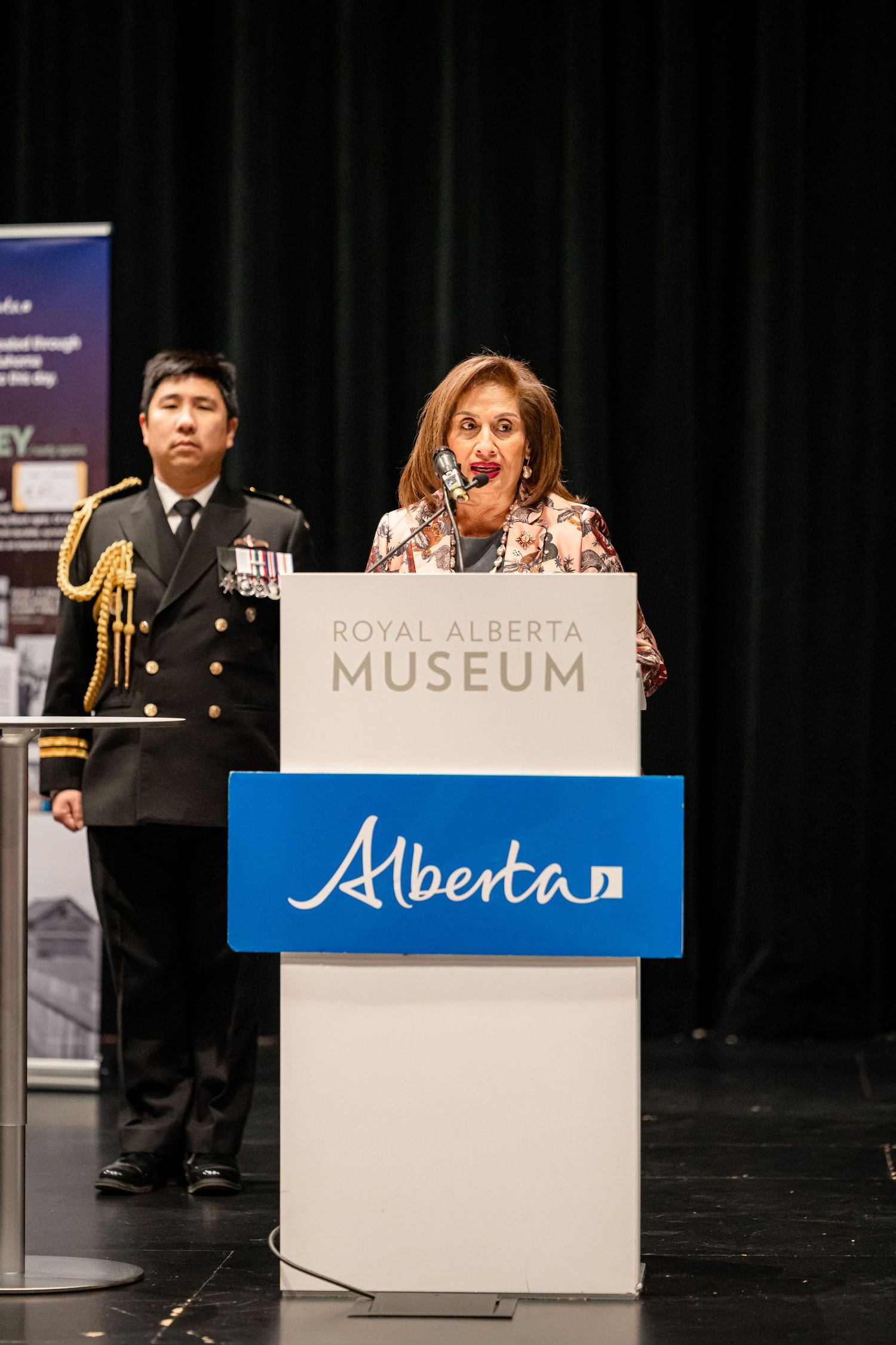Speaker at Royal Alberta Museum podium with military personnel standing nearby.