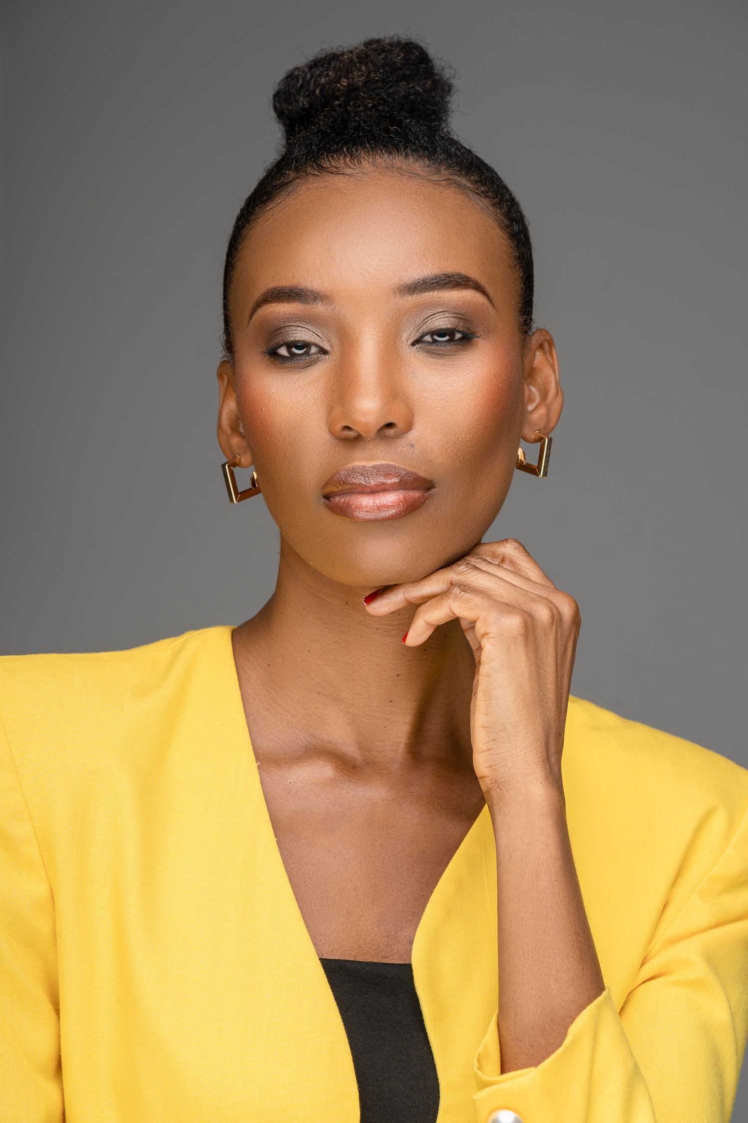 Professional portrait of a model in a bright yellow blazer with elegant makeup and hair styled in a bun against grey background.