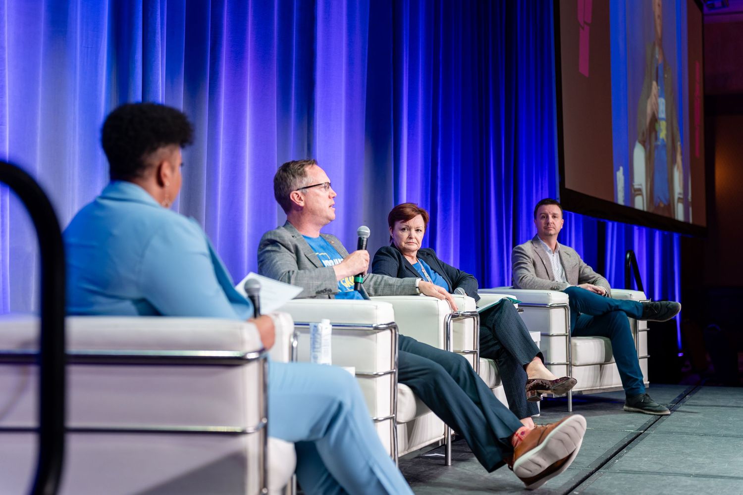 Conference panel discussion with speakers seated in white chairs on stage with blue lighting.