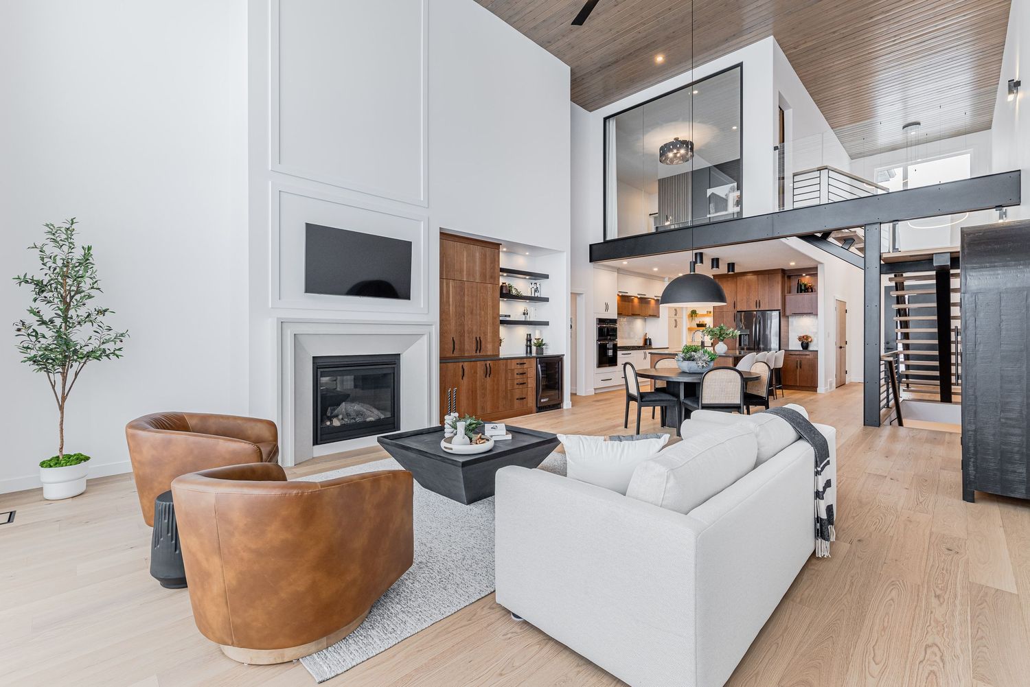 Modern open-concept living room with high ceilings, white sofa, leather chairs, fireplace, and mezzanine level above.