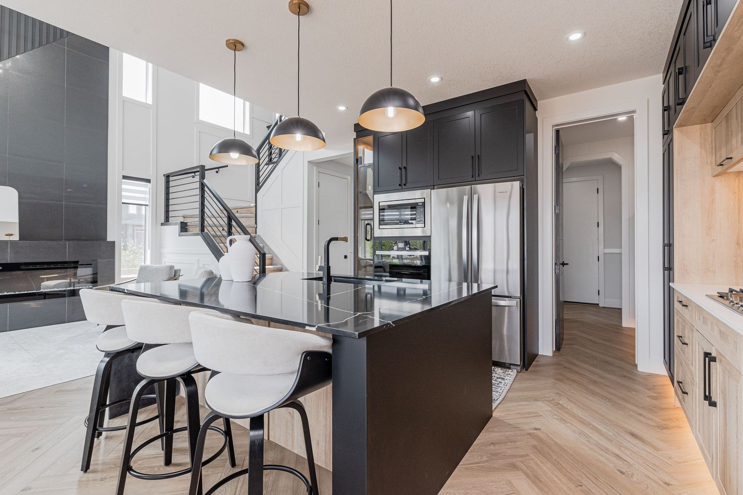 Modern kitchen with dark cabinetry, island seating, pendant lights, and stainless steel appliances on herringbone hardwood floors.