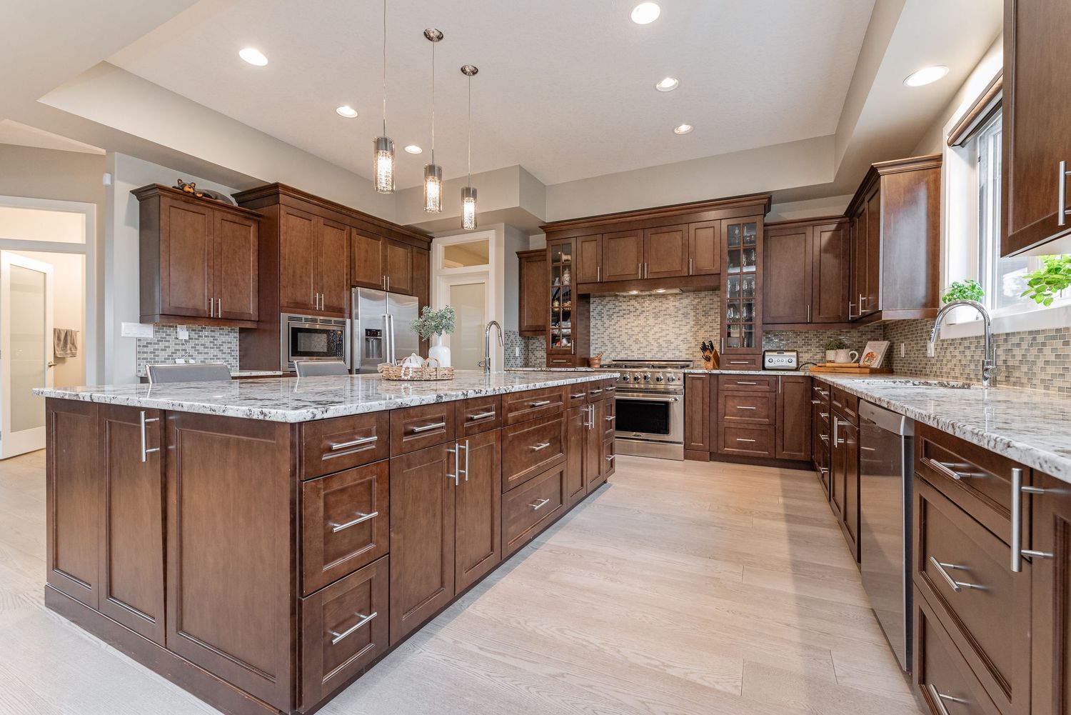 Spacious kitchen with dark wood cabinets, granite island, pendant lights, and stainless steel appliances on light hardwood floors.