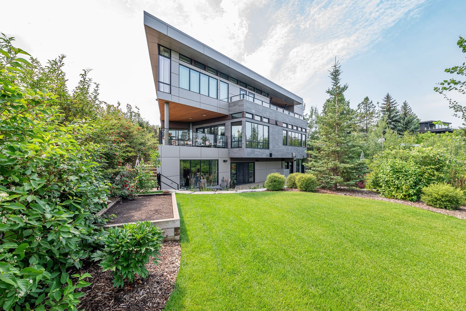 Modern multi-story home with large glass windows, wood accents, and a lush green lawn surrounded by mature trees.