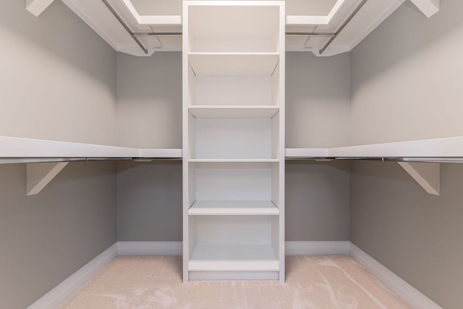 Empty walk-in closet with white shelving units, hanging rods, and neutral gray walls on light hardwood floors.