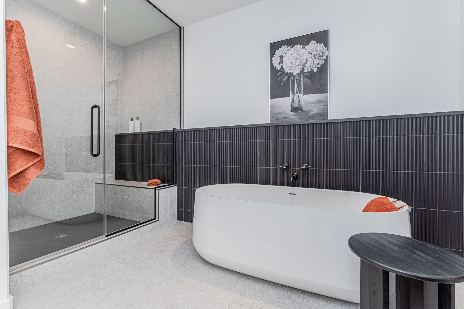 Modern bathroom with freestanding white tub, dark tile accent wall, glass shower, and orange towel accents.