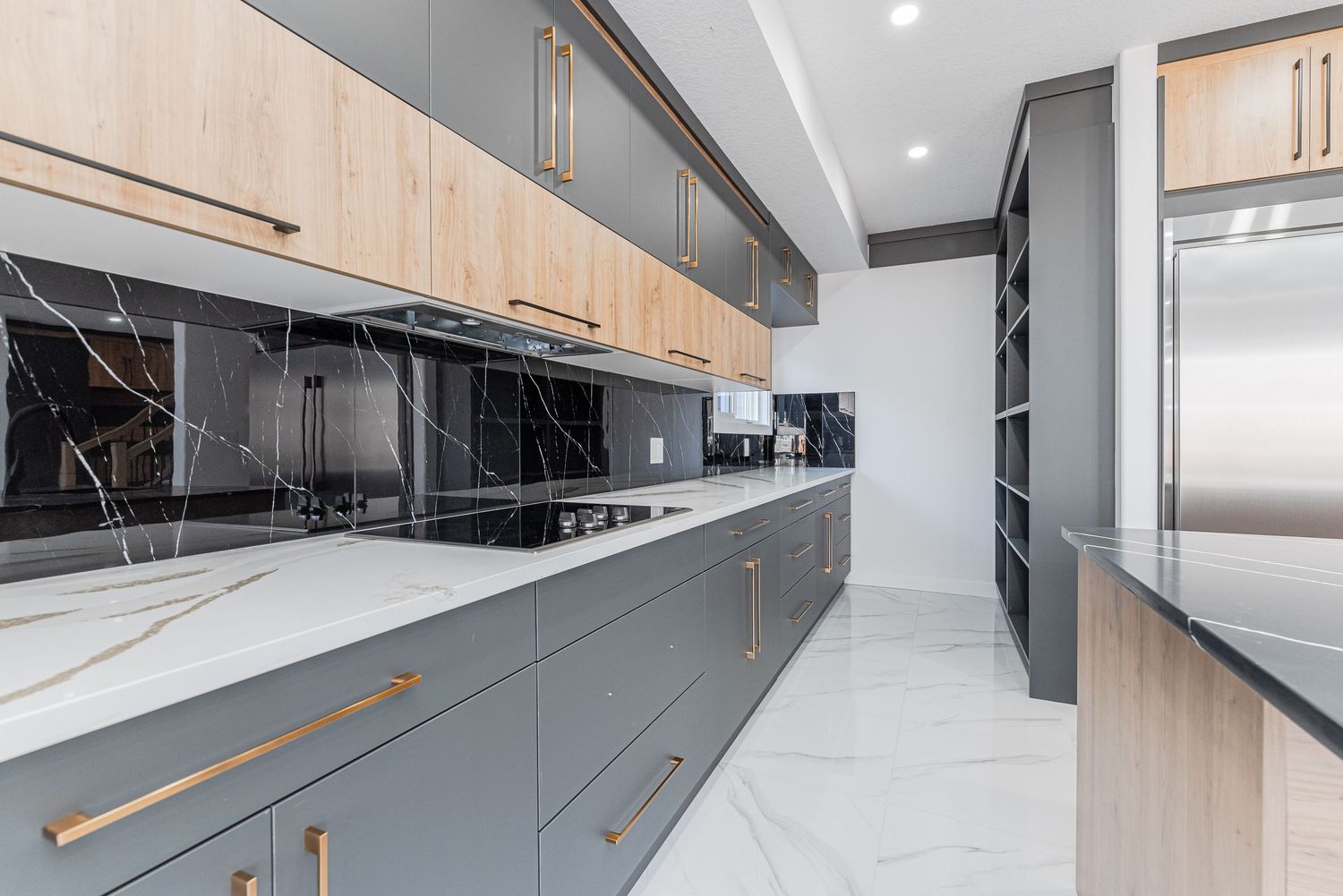 Modern kitchen with gray cabinets, wood accents, gold handles, marble countertops, and black marble backsplash.