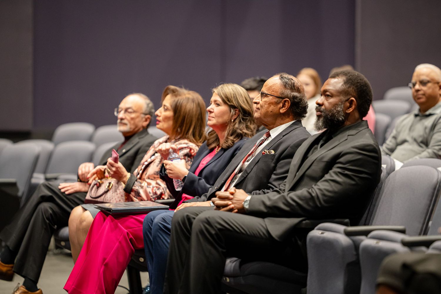 Audience members seated in rows of gray chairs watching a presentation intently.