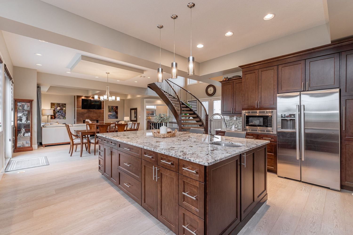 Spacious open-concept kitchen with dark wood cabinets, granite island, stainless steel appliances, and light hardwood floors.
