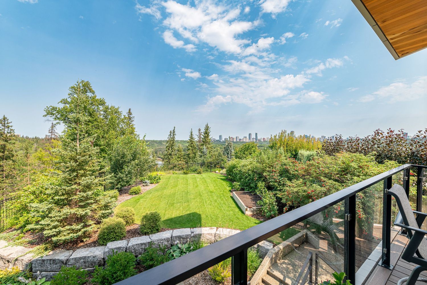 Balcony with glass railings overlooking a lush green garden and distant city skyline under a clear blue sky.