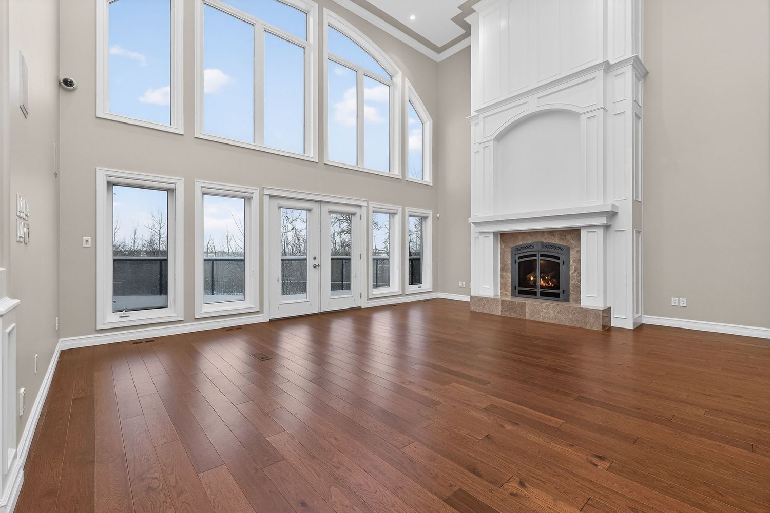 Spacious living room with hardwood floors, two-story windows, French doors, and an elegant white fireplace.