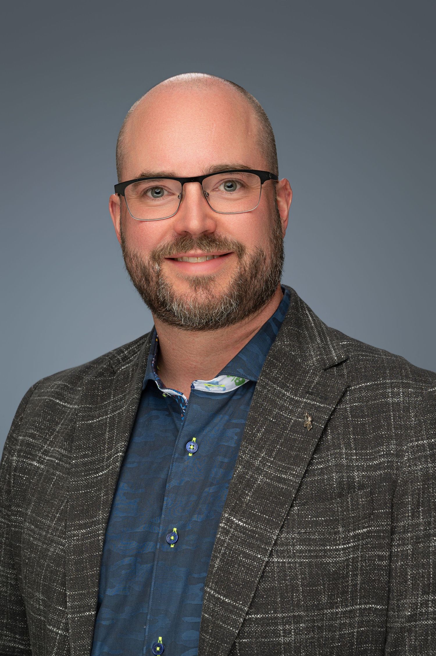 Professional headshot of person wearing glasses and plaid blazer with blue shirt.