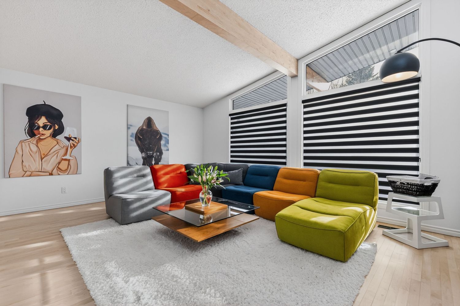 Modern living room with colorful modular sofa, wood beam ceiling, abstract wall art, and striped roller blinds on large windows.