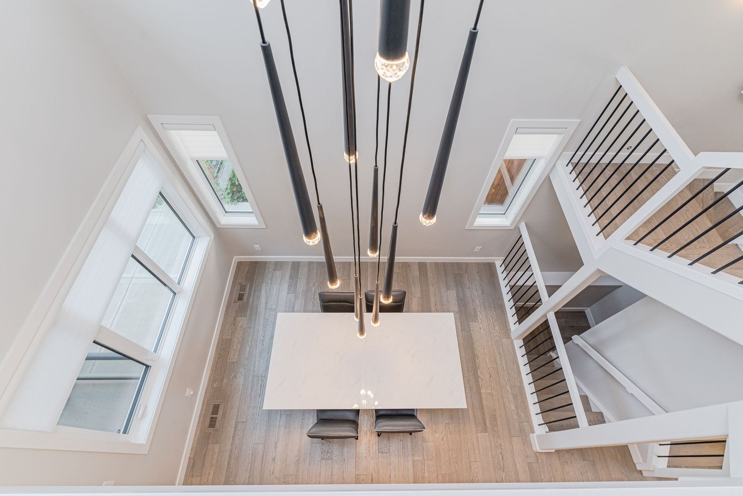 Aerial view of modern dining room with sleek pendant lighting, white table, and open staircase with light wood floors.