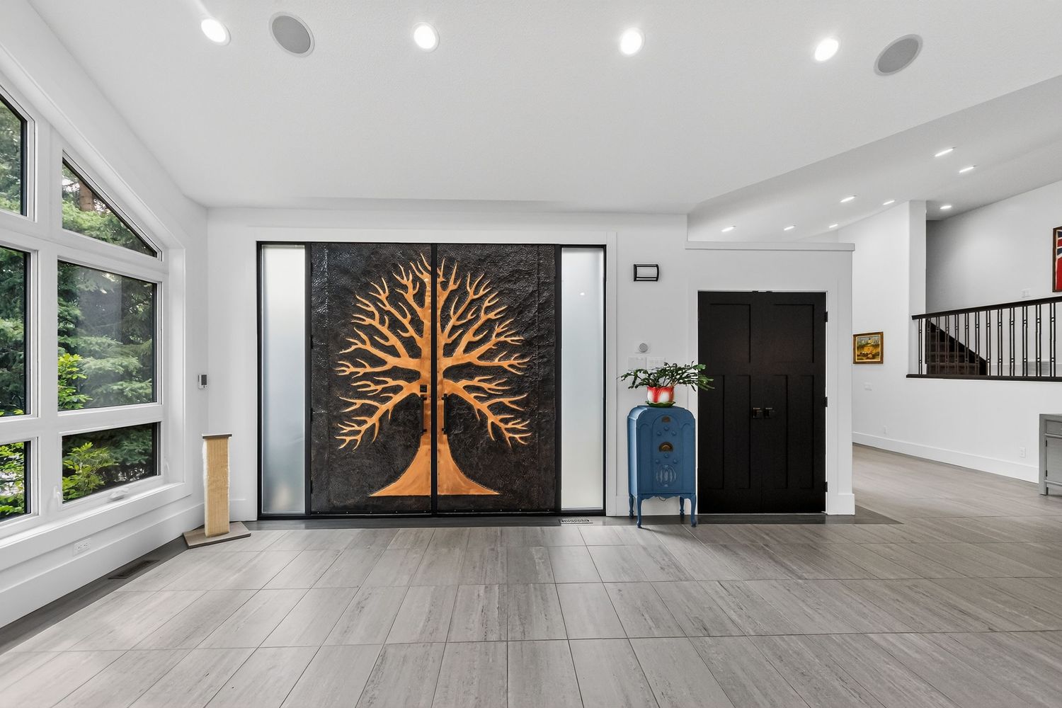 Modern foyer with large tree wall art, dark double doors, gray tile floors, and staircase in background.