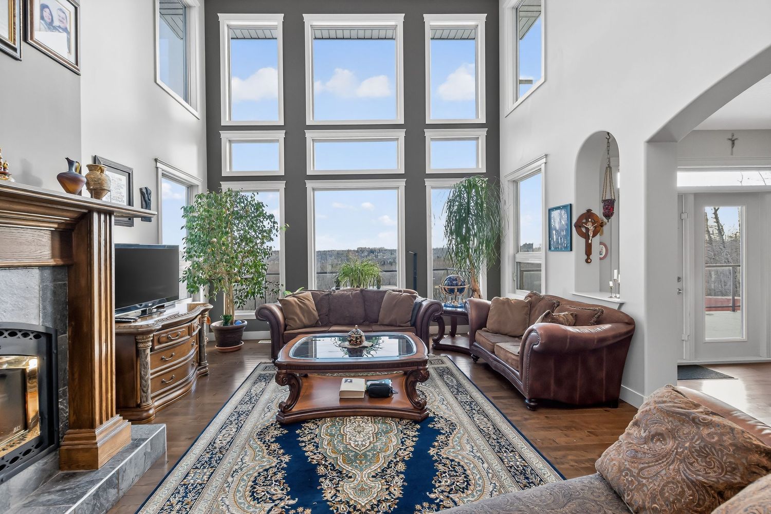 Elegant living room with soaring two-story windows, brown leather sofas, ornate rug, and a classic fireplace with hardwood floors.