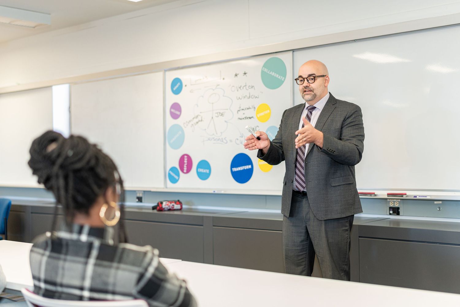 Professor giving presentation at whiteboard with colorful diagrams and charts in modern classroom setting.