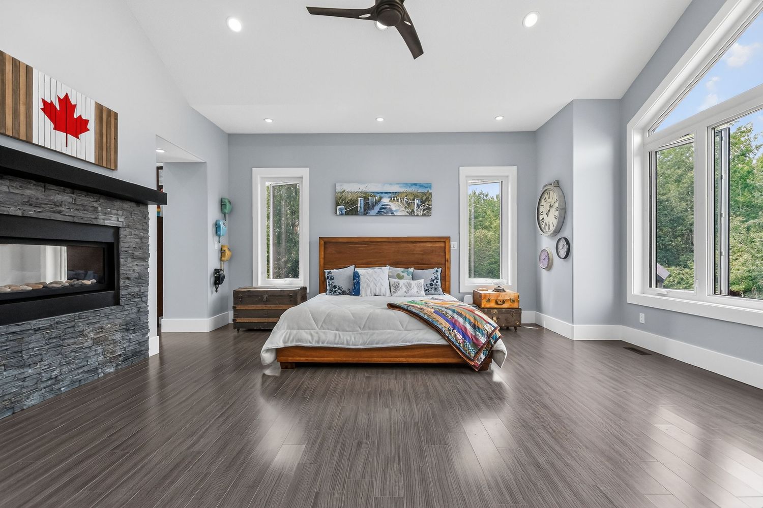 Spacious master bedroom with hardwood floors, stone fireplace, vaulted ceilings, ceiling fan, and large windows with natural light.