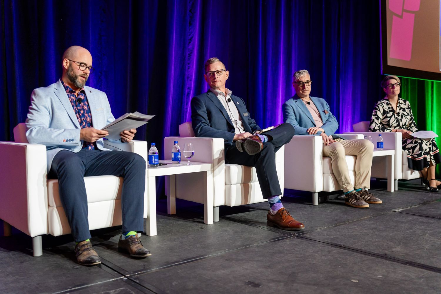Panel discussion with speakers seated on white chairs on stage at business conference.