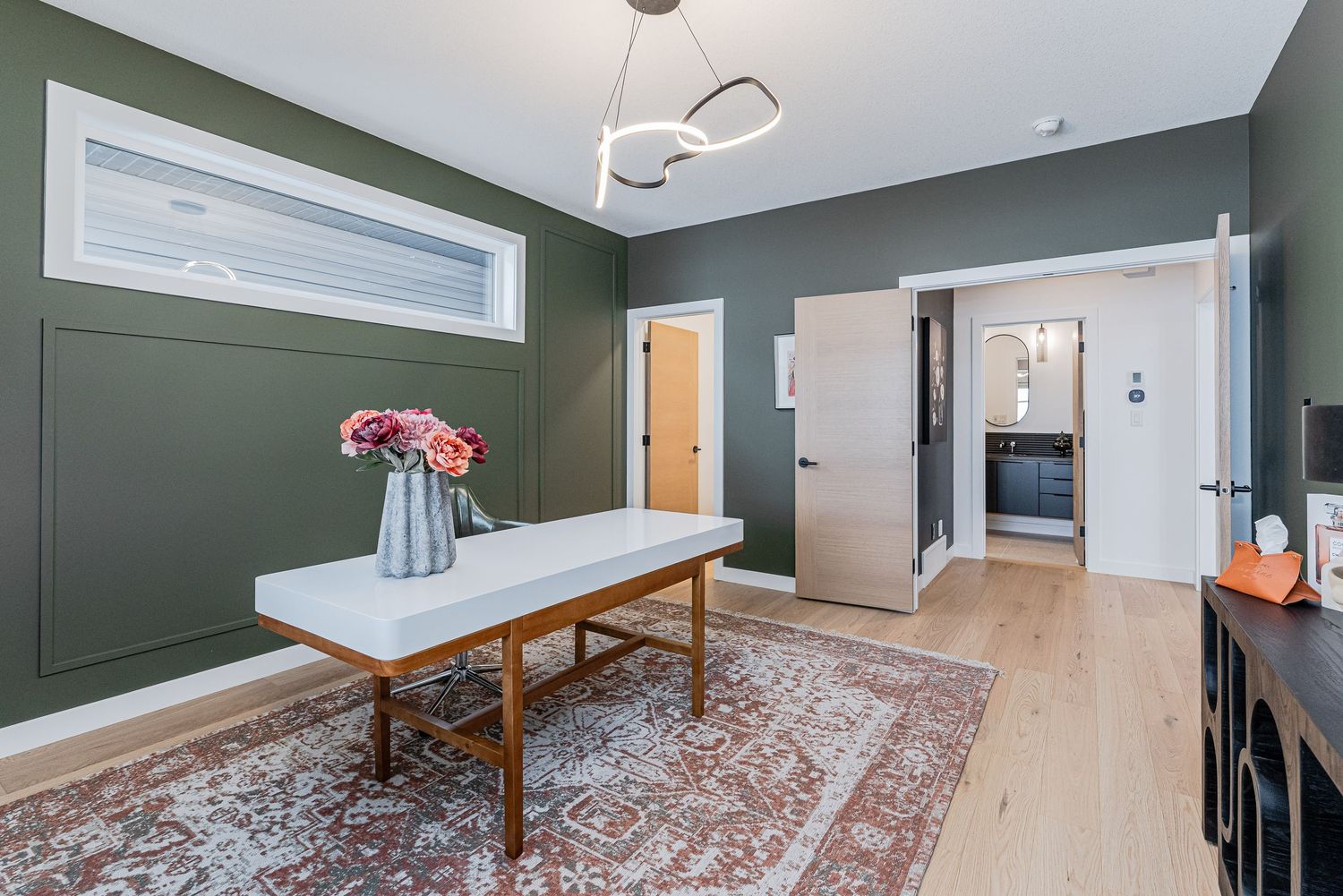 Modern home office with sage green walls, wooden desk, floral vase, patterned rug, and light hardwood floors.
