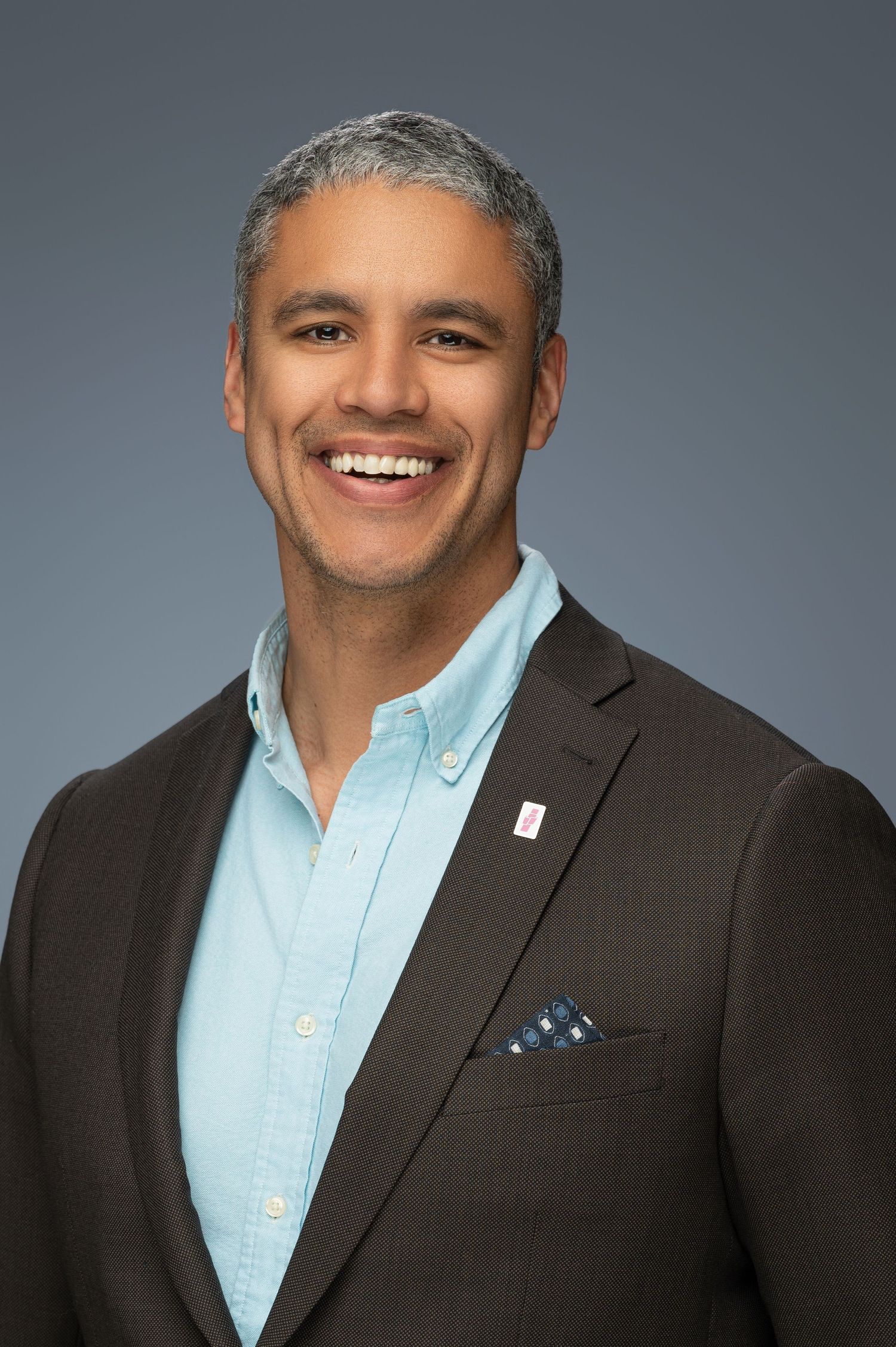 Professional portrait of person in dark suit and light blue shirt against gray background.