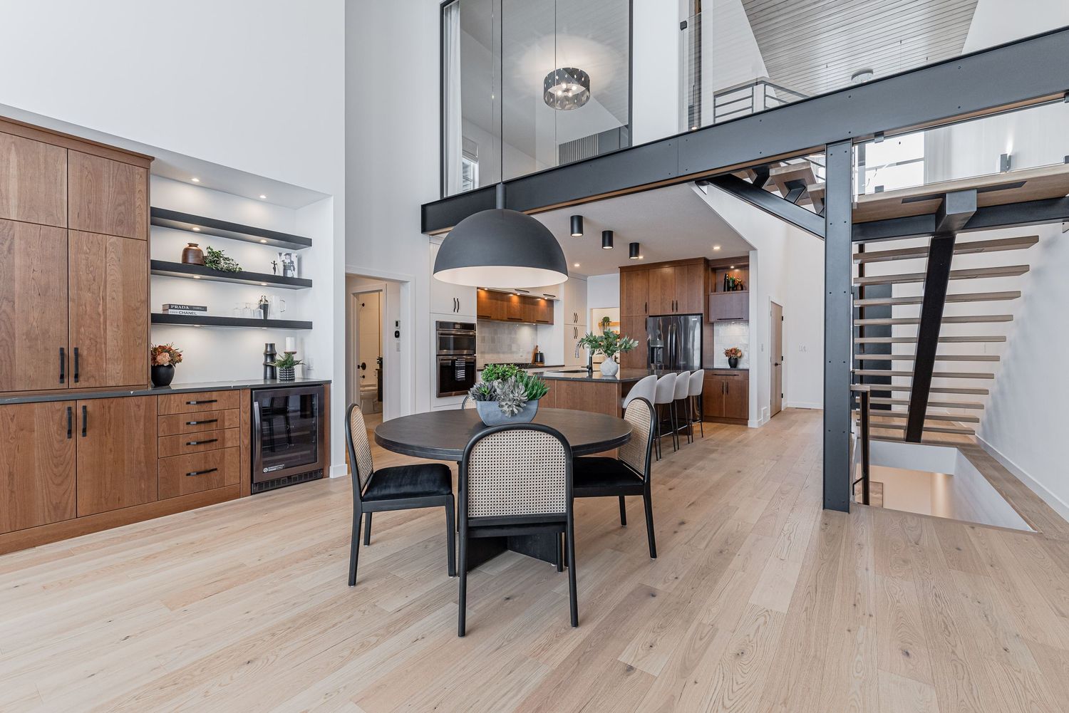 Modern open-concept dining area with light hardwood floors, black pendant light, wood cabinets, and open staircase.