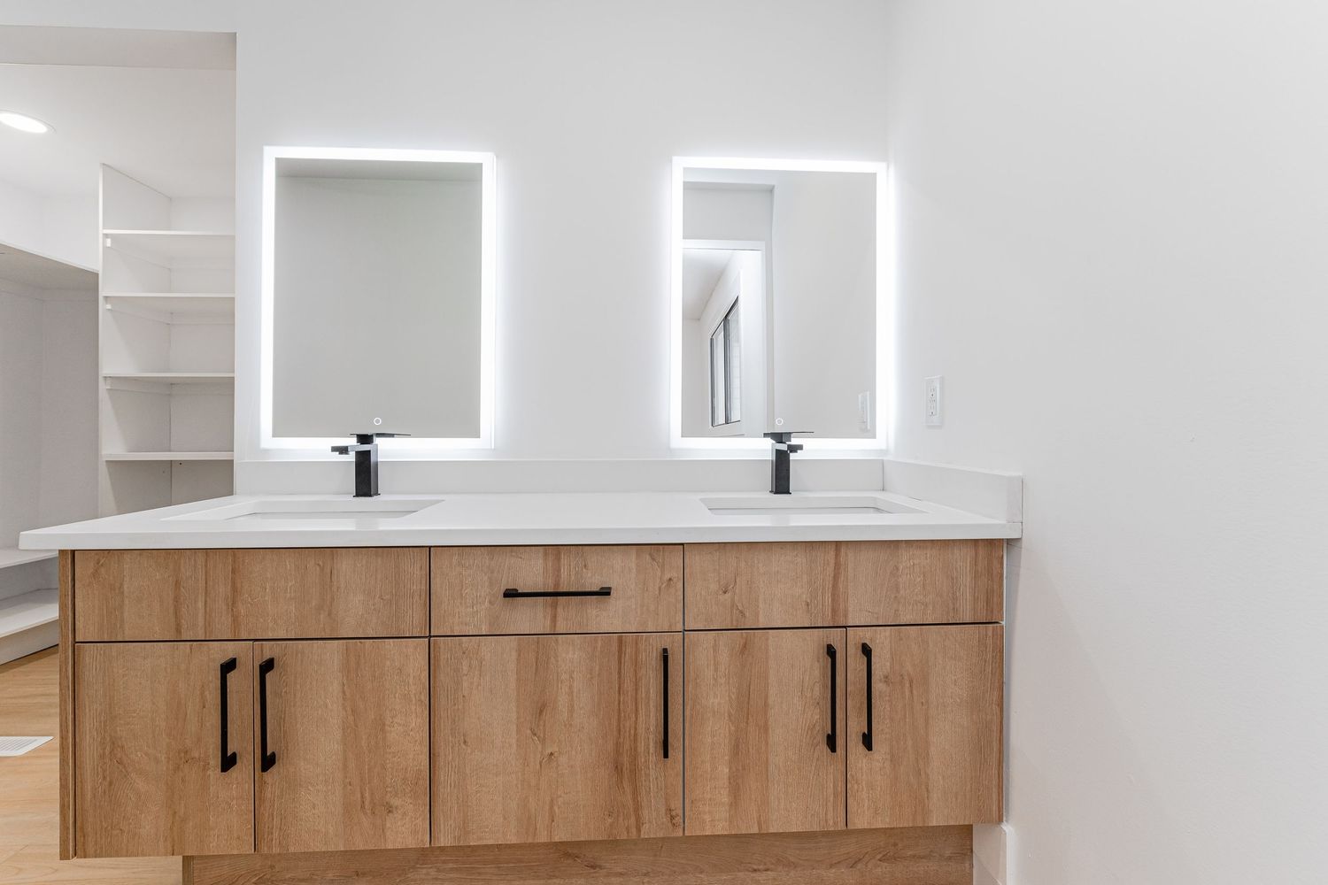 Modern double vanity with wood cabinets, black hardware, white countertop, and LED backlit mirrors in a bright bathroom.