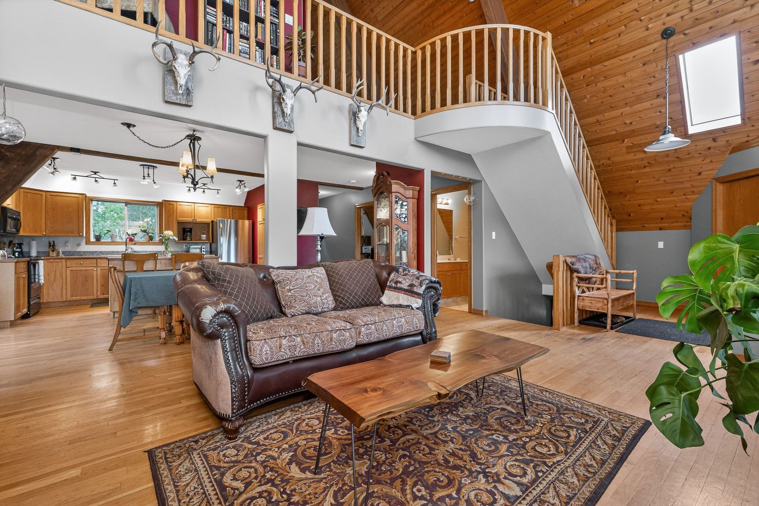 Spacious open-concept living room with hardwood floors, leather sofa, vaulted wood ceiling, staircase, and rustic decor.