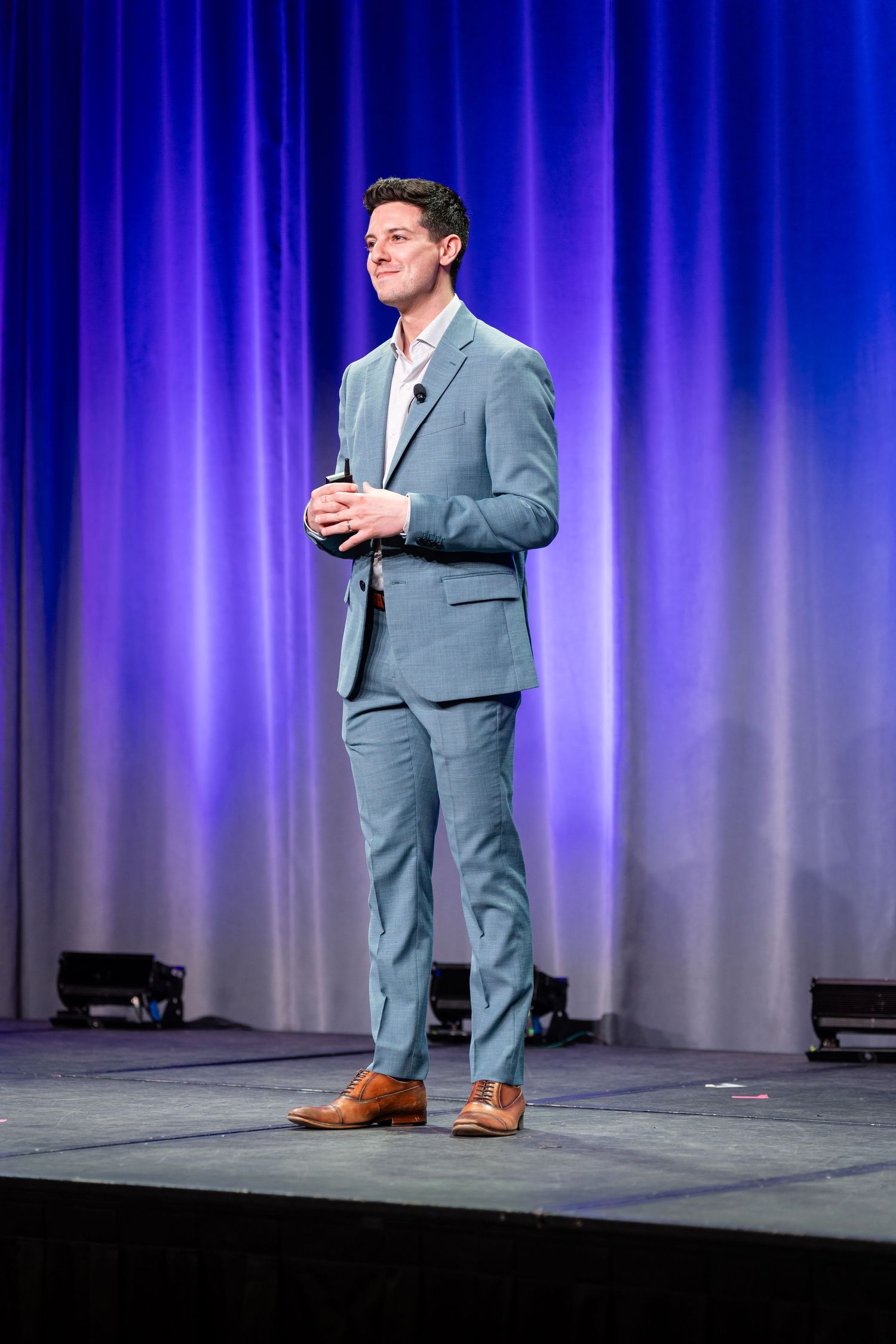Speaker presents on stage with blue curtain backdrop wearing grey suit.