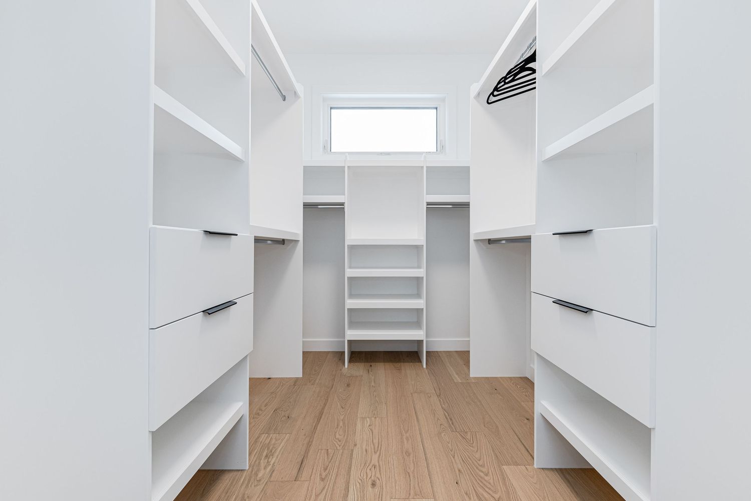 Bright white walk-in closet with built-in shelving, hanging rods, wood floors, and a small window for natural light.