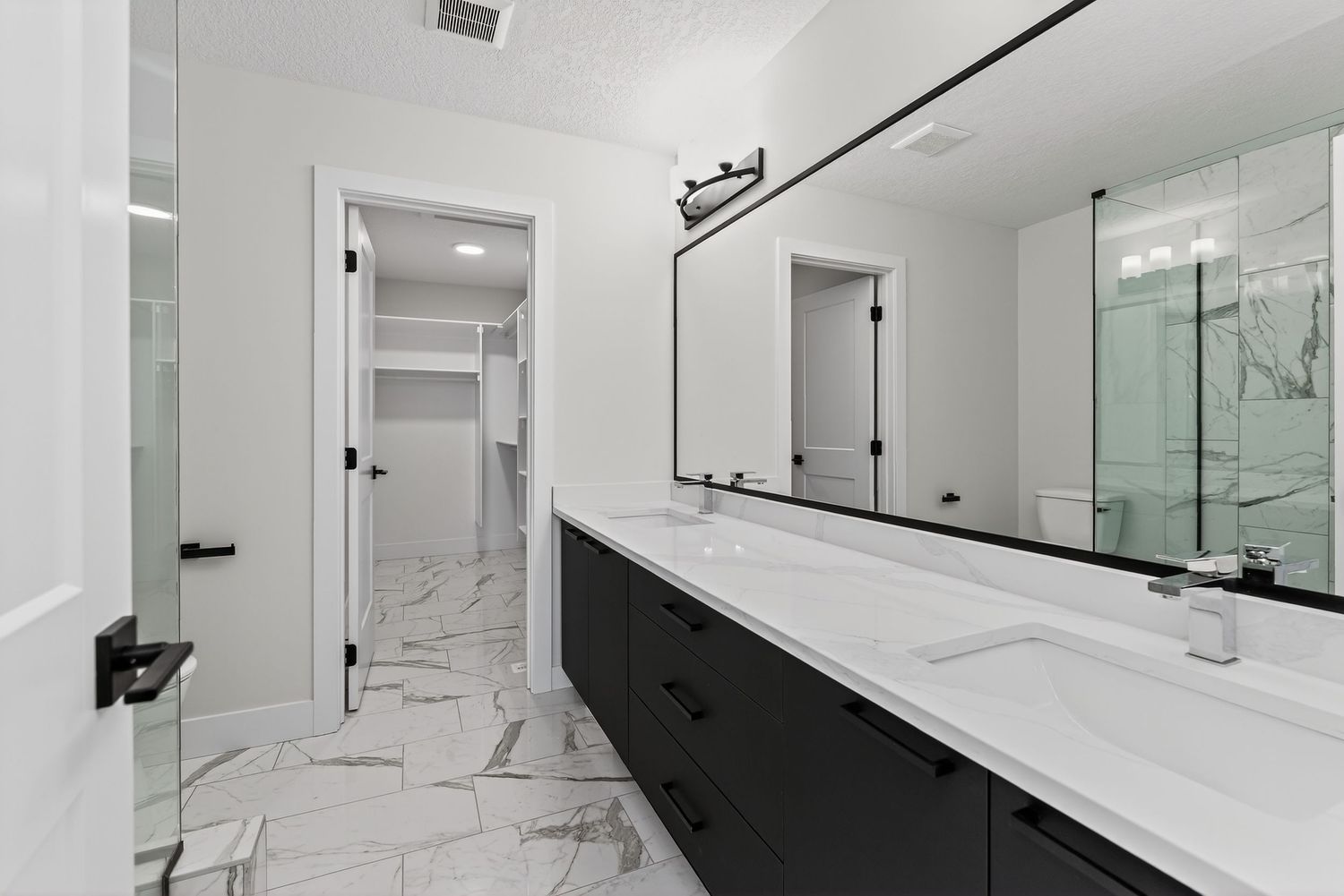Modern bathroom with dark vanity, dual sinks, white marble floors, and large mirror with black fixtures.