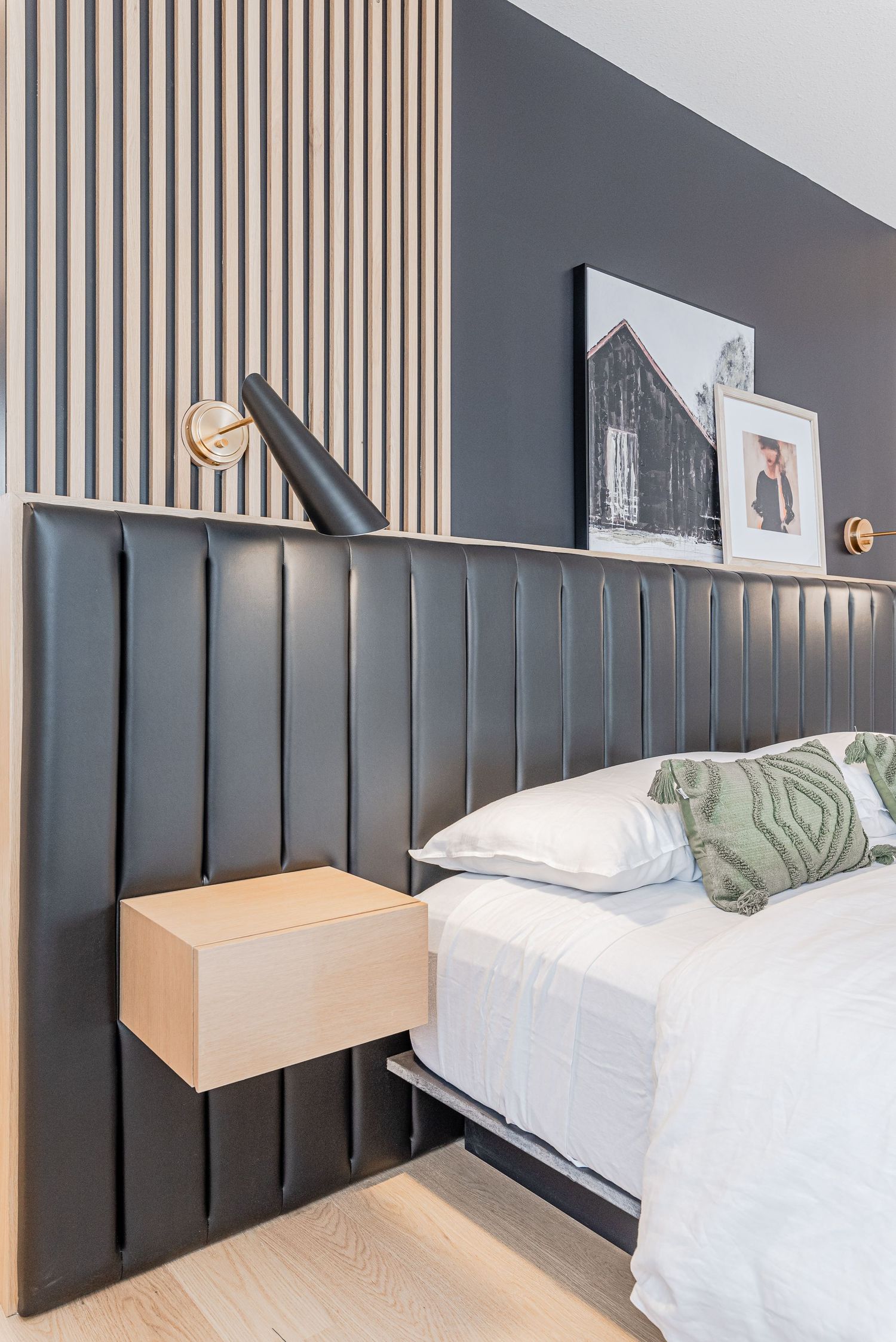 Modern bedroom with dark upholstered headboard, floating wood nightstand, brass wall sconce, and art shelf on gray accent wall.